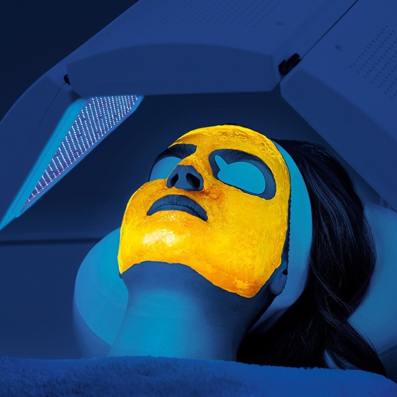 Woman with a bright yellow face mask receiving blue light therapy from an overhead machine.